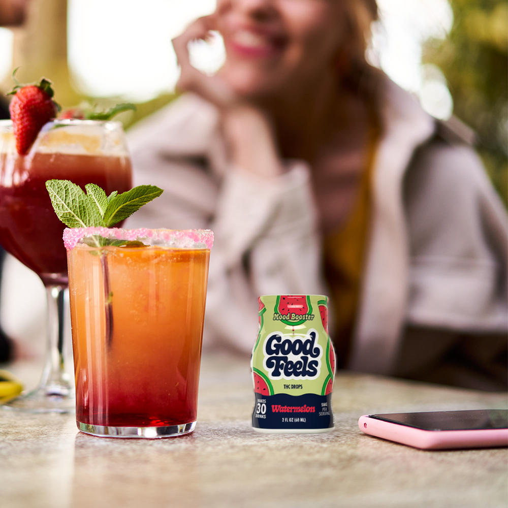 Two cocktails with a Good Feels Watermelon Beverage Enhancer on a table, with a blurred person in the background.
